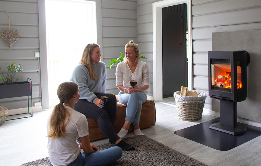 Kolme tyttöä istuvat ruskealla puffilla harmaassa olohuoneessa Jøtul F 167 -puukiukaan edessä